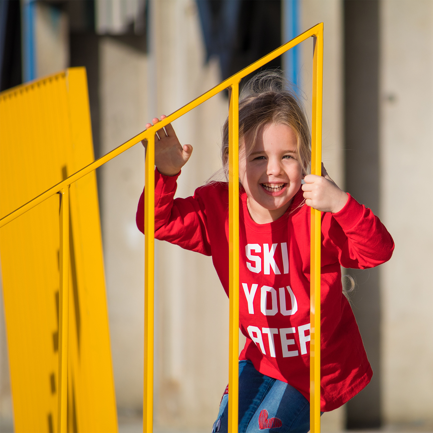 Blonde girl with red shirt with 'Ski je later' print by KMLeon behind yellow gate.