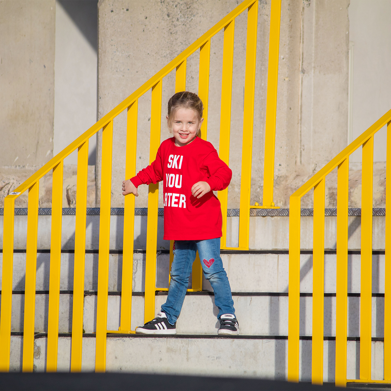 Blonde girl with red shirt with 'Ski je later' print by KMLeon next to yellow gate.