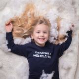 Blonde girl laying on snow with hair spread out and open arms, wearing navy shirt with 'I'm having a meltdown' print by KMLeon.