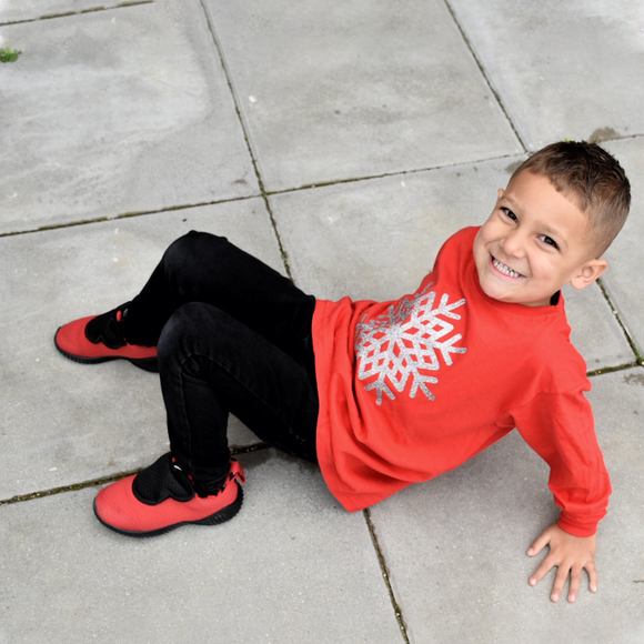 Boy with red shoes and red shirt with long sleeves with glitter snow star print sitting on concrete floor.