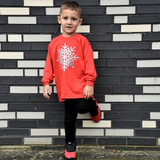 Boy with red shoes and red shirt with long sleeves with glitter snow star print sitting in front of black and white brick wall.
