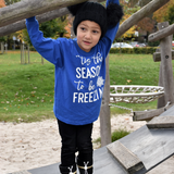 Boy at the playground wearing black hat with pompoms and blue shirt with long sleeves with ''tis the season to be freezin'' print by KMLeon.