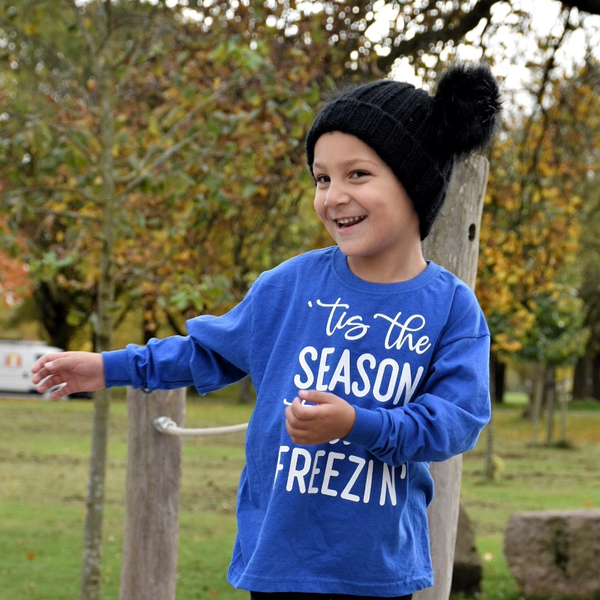Happy boy in nature wearing black hat with pompoms and blue shirt with long sleeves with ''tis the season to be freezin'' print by KMLeon.
