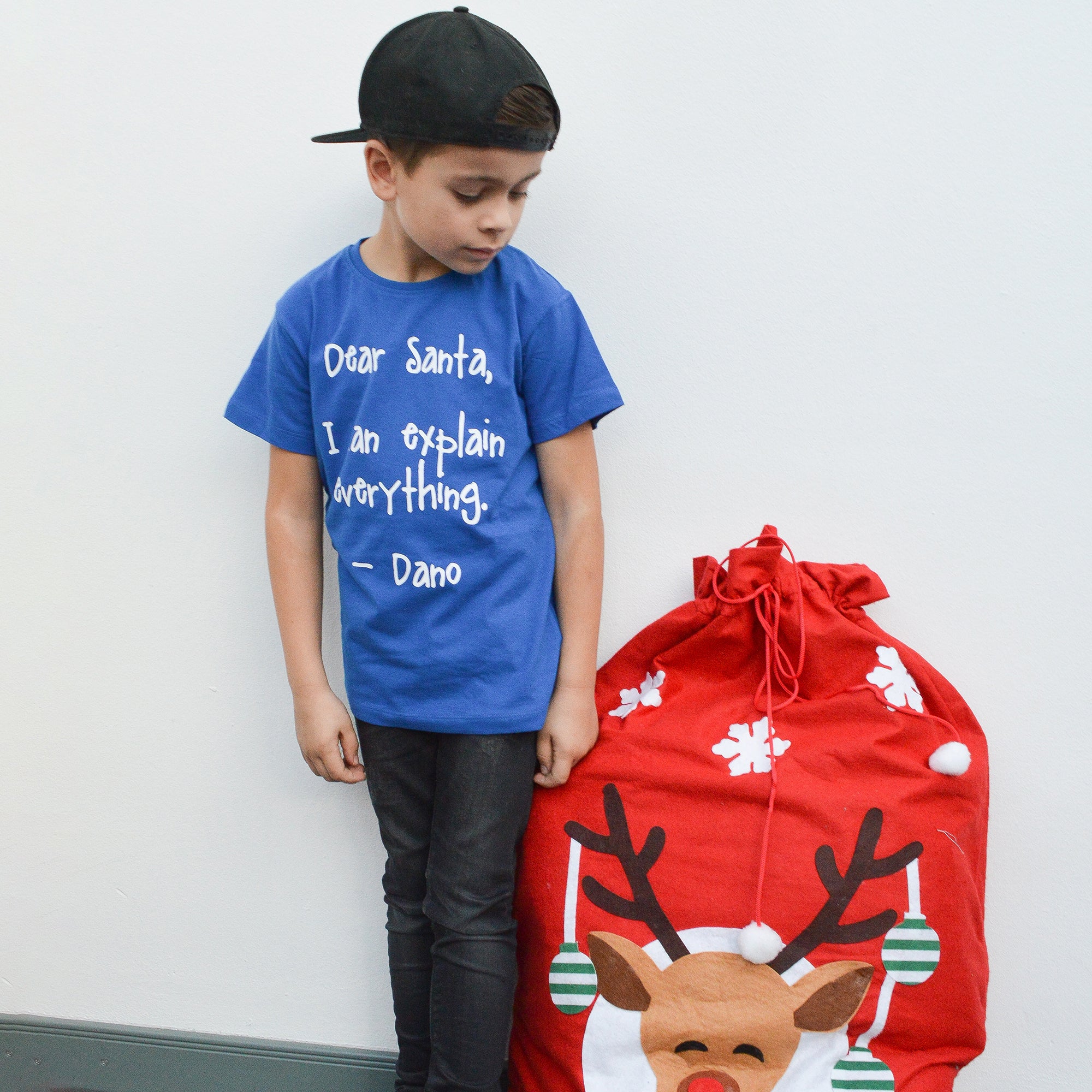 Boy with black cap and blue shirt with 'Santa, I can explain everything' shirt by KMLeon, looking at red christmas bag.
