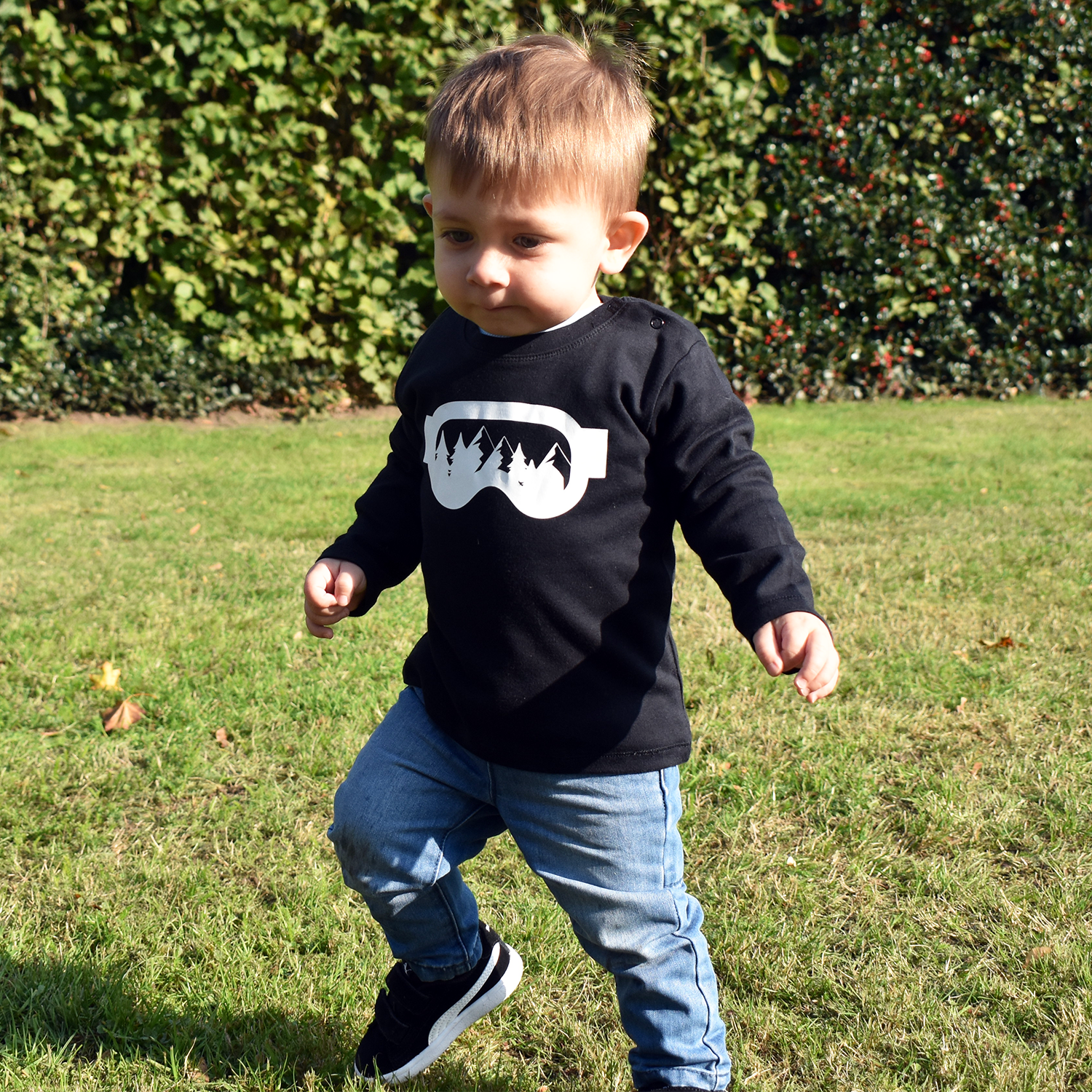 Little boy wearing black shirt with longs sleeves withh ski goggles print by KMLeon walking in park.