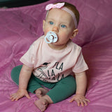 Baby girl with blue eyes, wearing a ppink headband and pink shirt, with 'Fla la la la la mingo' print by KMLeon looking to the side.
