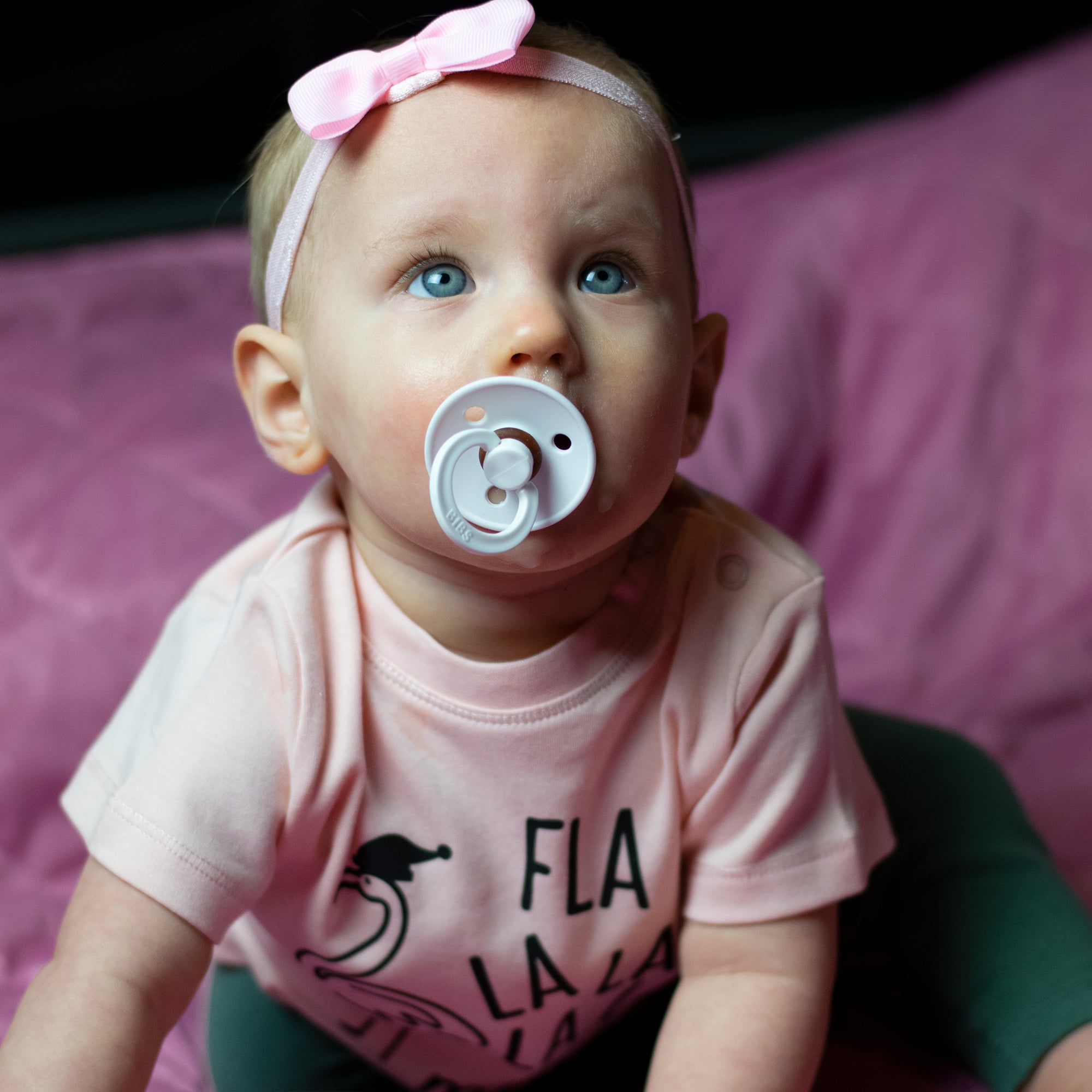 Baby girl with blue eyes, wearing a ppink headband and pink shirt, with 'Fla la la la la mingo' print by KMLeon looking up with pacifier.