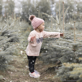 Blonde girl with pink beanie wearing pink shirt with long sleeves with glitter snow flake glasses print by KMLeon, grabbing christmas tree.