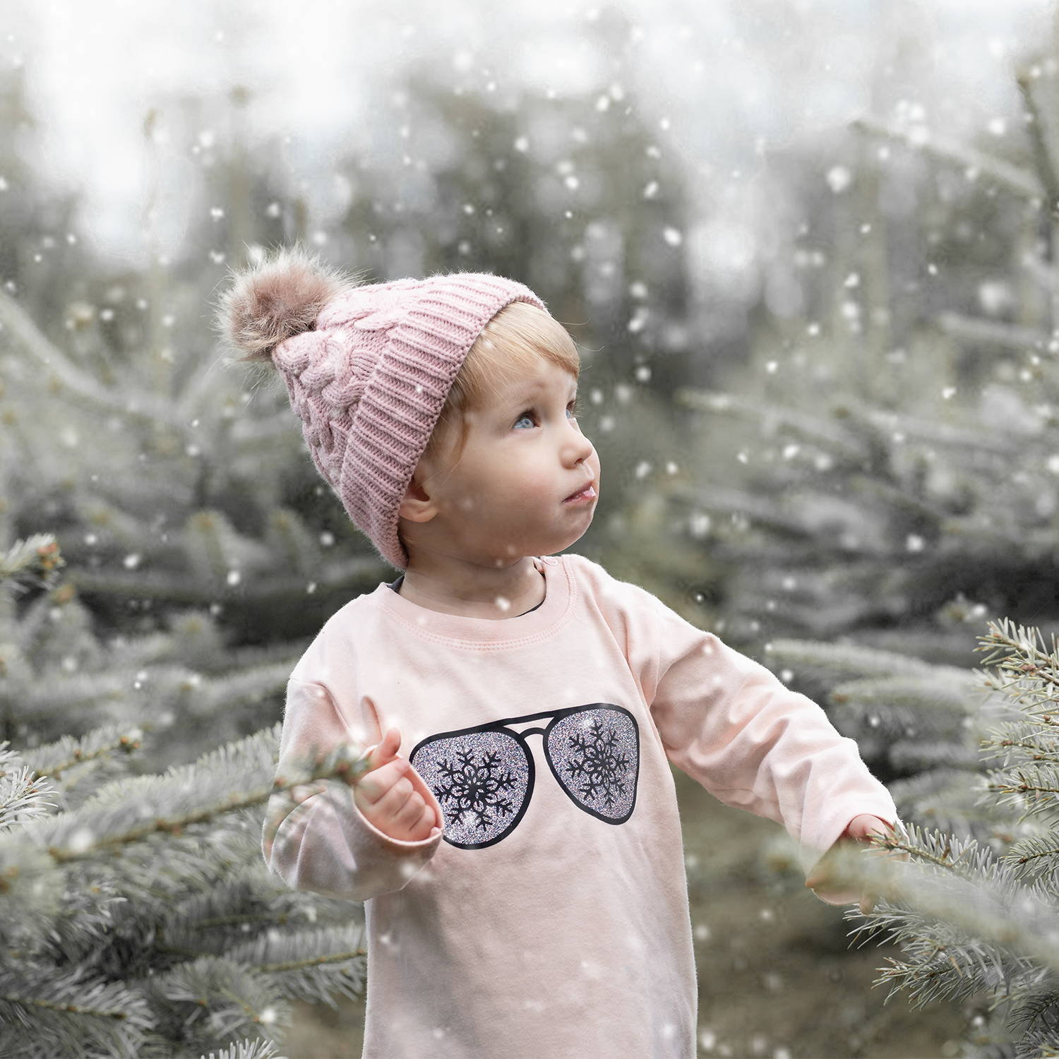 Blonde girl with pink beanie wearing pink shirt with long sleeves with glitter snow flake glasses print by KMLeon, looking at snow.