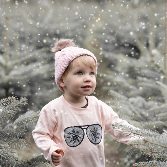 Blonde girl with pink beanie wearing pink shirt with long sleeves with glitter snow flake glasses print by KMLeon, looking at snow.