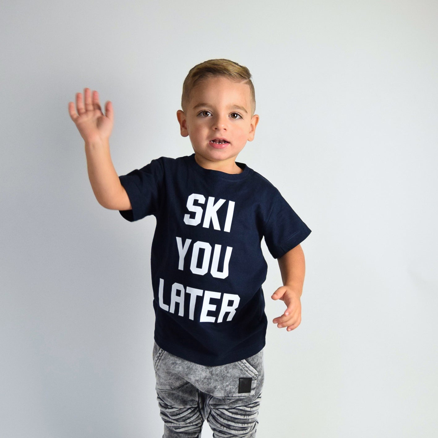 Boy wearing navy shirt with 'Ski je later' print by KMLeon, waving.