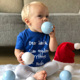 Blonde boy with blue shirt with 'Santa, I can eplain everything' print by KMLeon, putting christmas ornament in mouth.