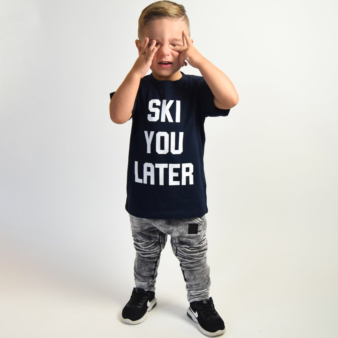 Boy wearing navy shirt with 'Ski je later' print by KMLeon, holding hands before eyes.