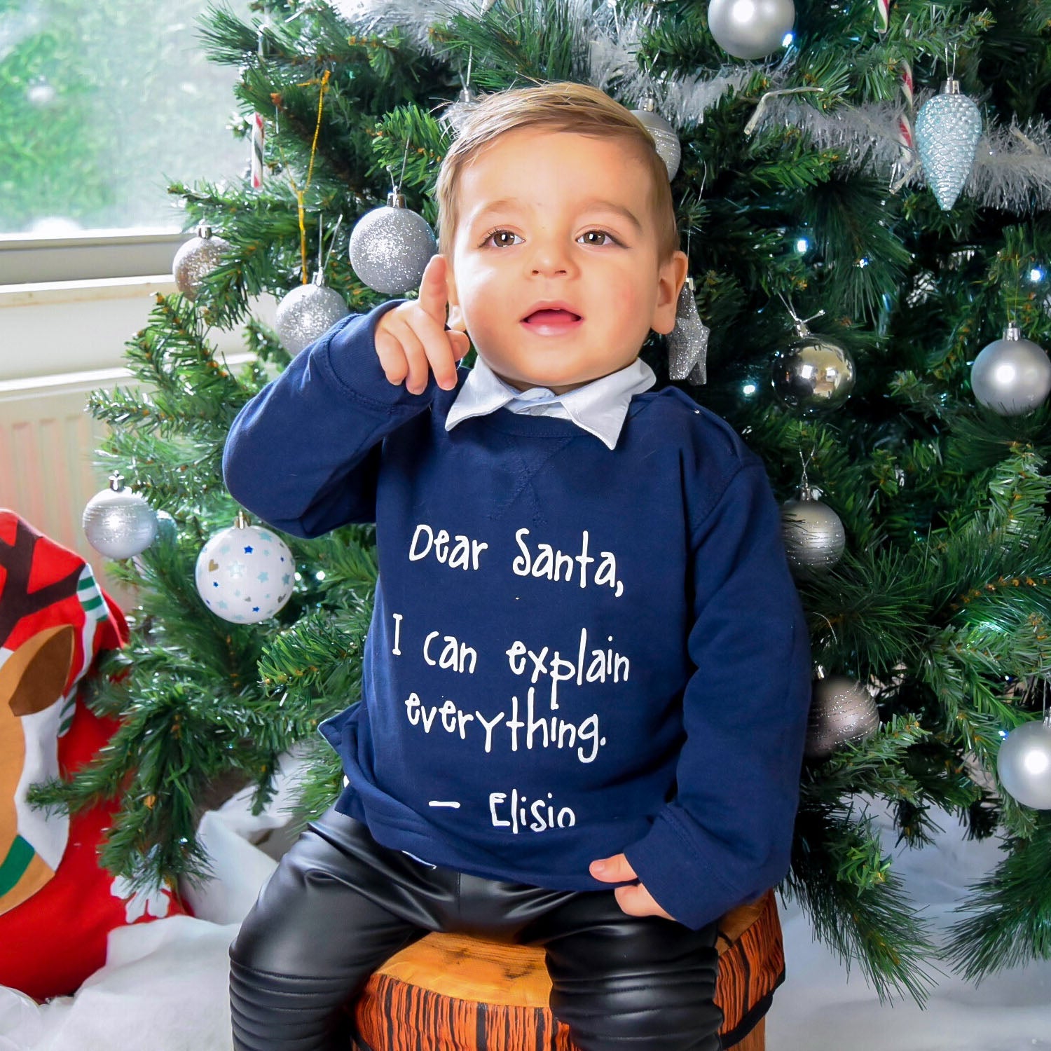 Boy with Navy sweater with 'Santa, I can explain everything' print by KMLeon in front of christmas tree, pointing his finger.