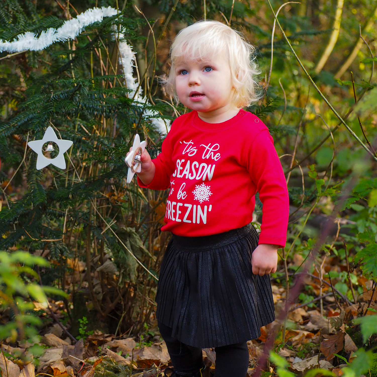 Blonde girl in forest holding white star wearing black skirt and red shirt with long sleeves with ''tis the season to be freezin'' print by KMLeon.