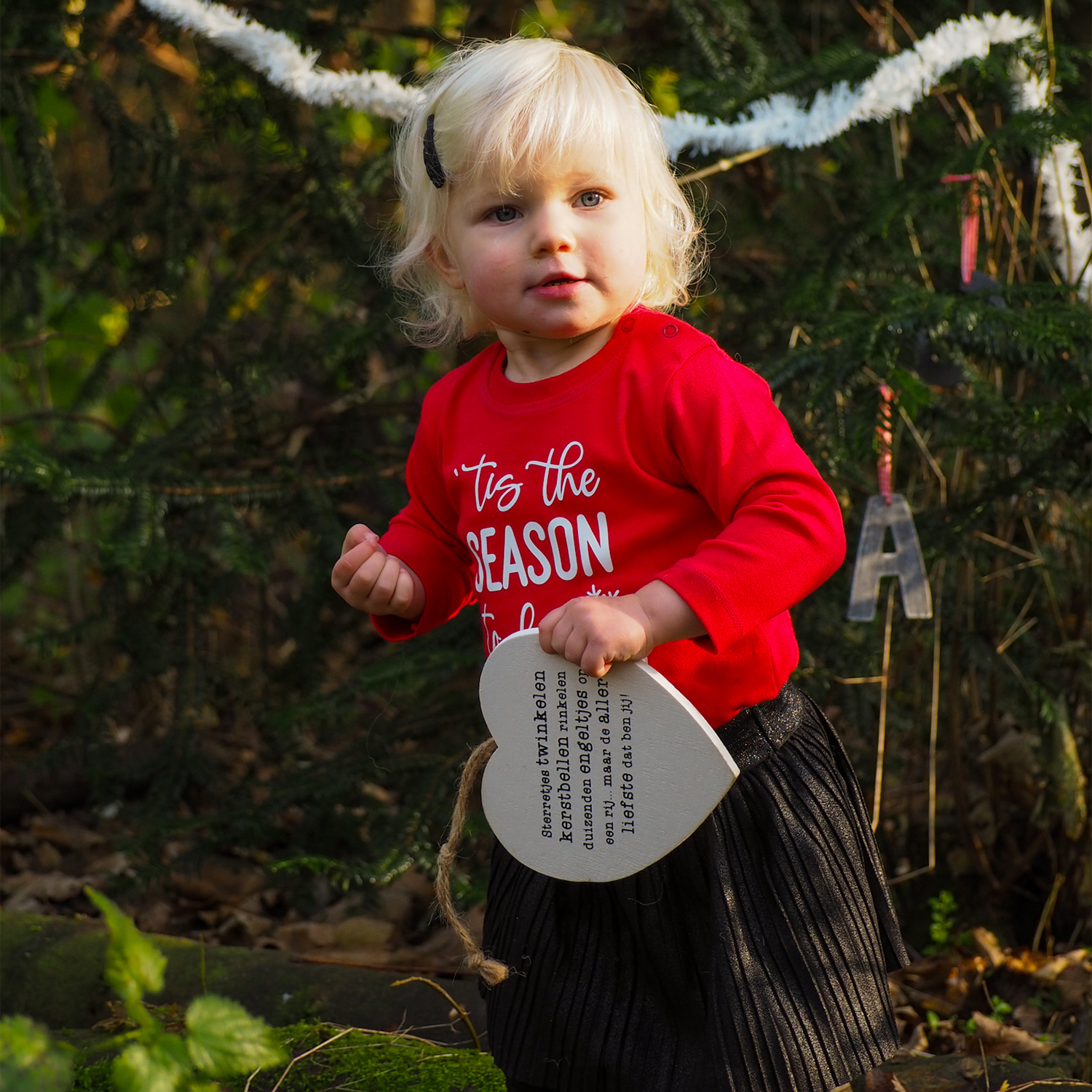 Blonde girl in forest holding wooden heart wearing black skirt and red shirt with long sleeves with ''tis the season to be freezin'' print by KMLeon.