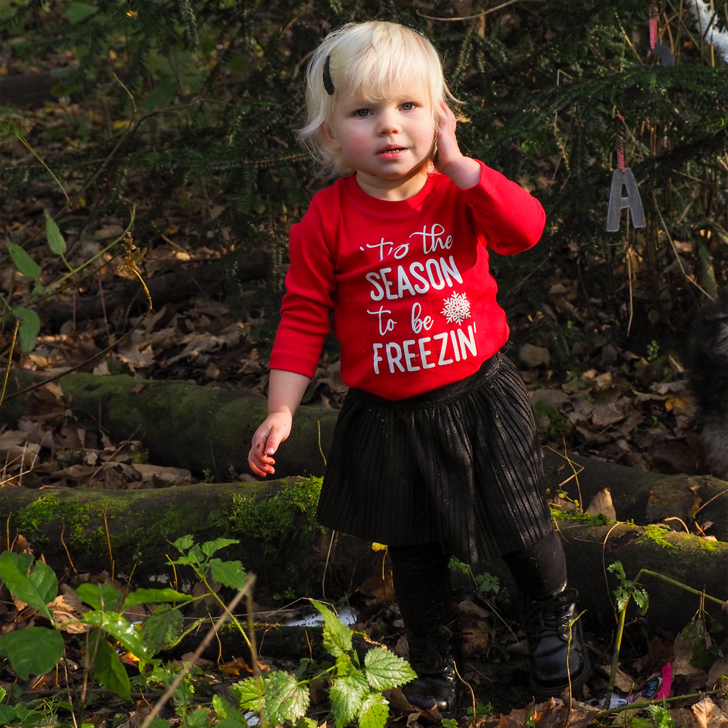 Blonde girl in forest with hand to ear wearing black skirt and red shirt with long sleeves with ''tis the season to be freezin'' print by KMLeon.