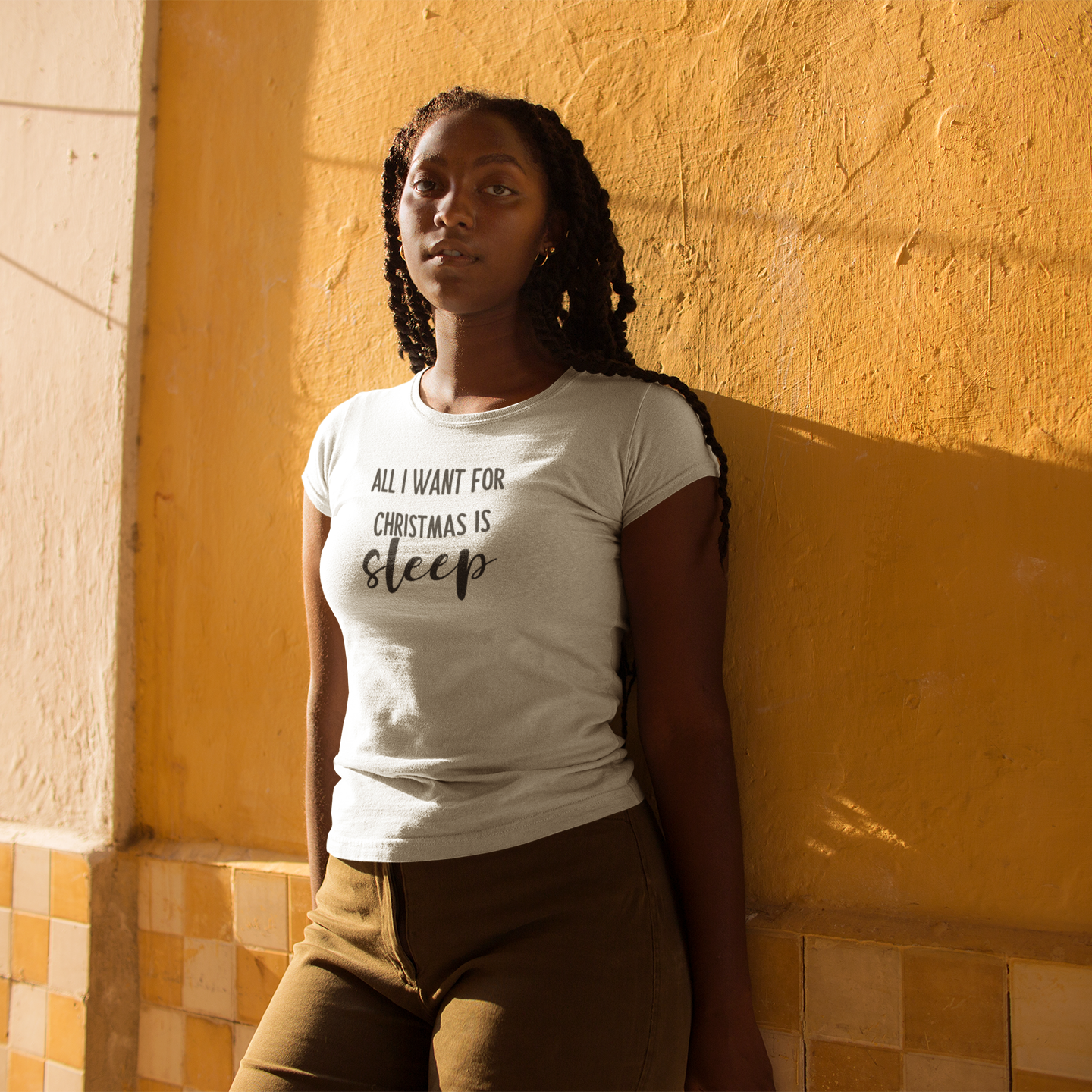 Black woman with braids in front of yellow wall, wearing white shirt with 'All I want for Christmas is sleep' print by KMLeon.