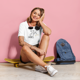 Smiling tattooed woman on yellow skateboard wearing white shirt with 'All the jingle ladies' print by KMLeon, in front of pink wall.