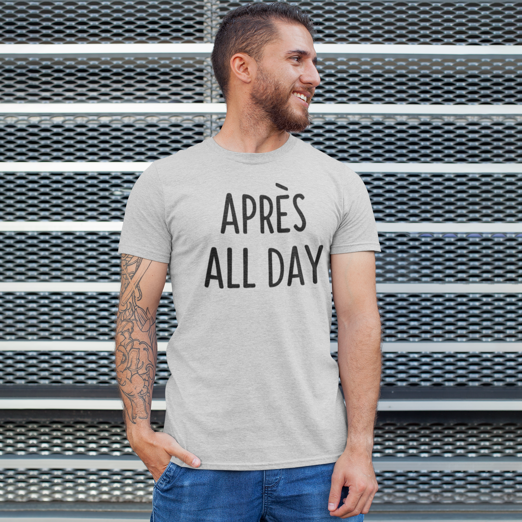 Man with beard and tattoo wearing heather grey shirt with 'Après all day' print by KMLeon, looking to the side.