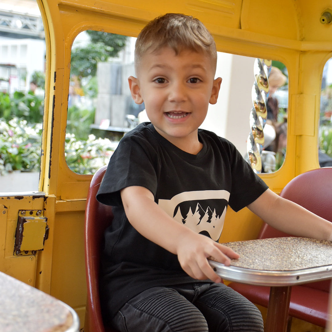 Young boy wearing black shirt with ski goggles print by KMLeon sitting in yellow bus.
