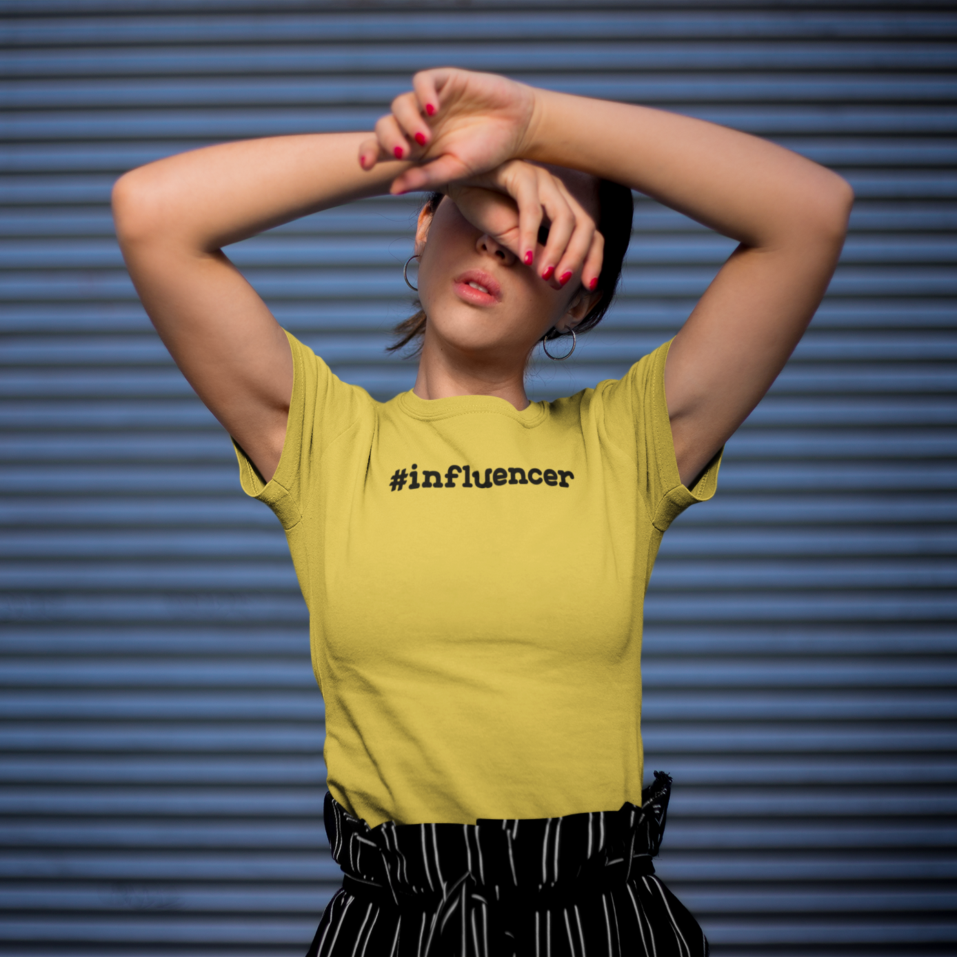 Women with yellow shirt with '#influencer' shirt by KMLeon against blue background, with arms in front of face.