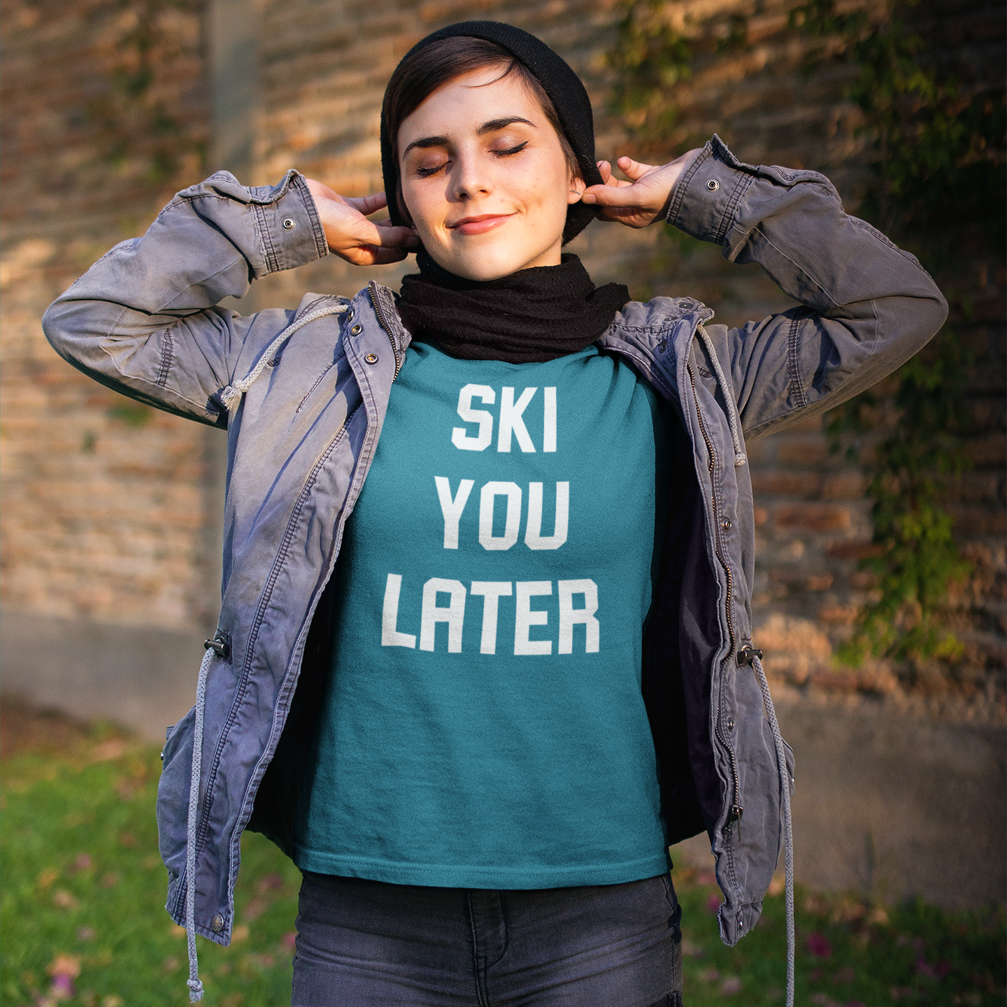 Women with aqua shirt, with 'Ski you later print' by KMLeon putting on black hat, with eyes closed.